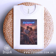 Футболка не ідеальний але твій. Біла футболка, складена на плетеному кошику, з принтом пари на фоні вечірнього освітлення.