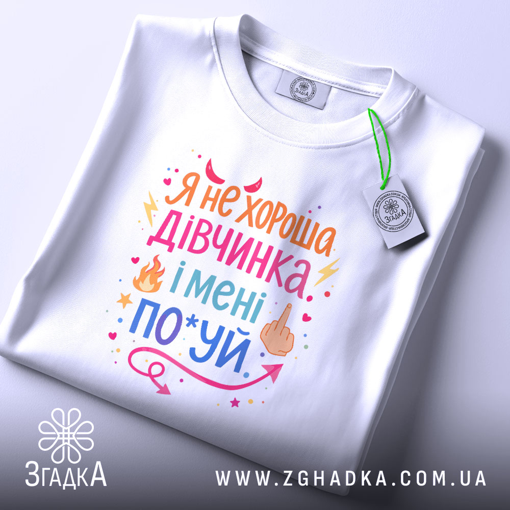 Футболка не хороша дівчинка. Біла футболка, складена на поверхні, з видимим принтом. Зображена з верхнього ракурсу. Фон світлий, з легким градієнтом. Принт кольоровий, з текстом та графічними елементами.