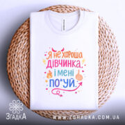 Футболка не хороша дівчинка. Біла футболка, складена на плетеній основі, зображена з верхнього ракурсу. Фон світлий. Принт кольоровий, з текстом та графічними елементами, розташований по центру.