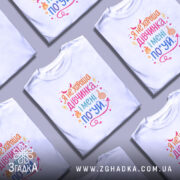 Футболка не хороша дівчинка. Біла футболка з кольоровим принтом, зображена з фронтального ракурсу на манекені. Фон нейтральний, з легким градієнтом. Принт містить текст з яскравими кольорами, символами вогню та жестом середнього пальця.