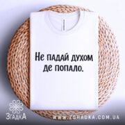 Футболка не падай духом, білого кольору, складена на плетеній кошику, фон світло-сірий, принт чорного кольору з текстом 'Не падай духом де попало.'