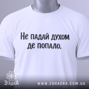 Футболка не падай духом, білого кольору, складена на поверхні, фон світло-сірий, принт чорного кольору з текстом 'Не падай духом де попало.', на комірці видно етикетку.