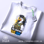 Футболка не святий але свій, біла, зображена зверху, на столі, фон світлий, принт з птахом у військовій формі, з написом 'НЕ СВЯТИЙ АЛЕ СВІЙ', прикріплена етикетка з зеленим шнурком.