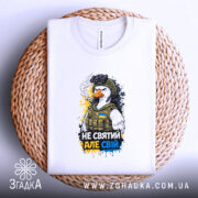 Футболка не святий але свій, біла, зображена зверху на плетеній основі, фон світлий, принт з птахом у військовій формі, з написом 'НЕ СВЯТИЙ АЛЕ СВІЙ', деталі принта яскраві та виразні.