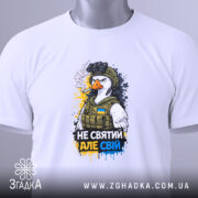 Футболка не святий але свій, біла, зображена зверху, на столі, фон світлий, принт з птахом у військовій формі, з написом 'НЕ СВЯТИЙ АЛЕ СВІЙ', прикріплена етикетка з зеленим шнурком.