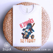 Футболка не зупиняйся, біла, зображена зверху на плетеній основі, фон білий, принт з малюнком ведмедя на триколісному велосипеді з червоним текстом 'НЕ ЗУПИНЯЙСЯ' та 'ГАЗ У ПІДЛОГУ'.