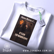Футболка нема батога. Біла футболка, складена на поверхні, з видимим етикеткою. Зображення ведмедя з пряником на фоні світло-сірого кольору. Принт з текстом на українській мові.