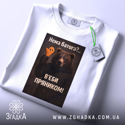 Футболка нема батога. Біла футболка, складена на поверхні, з видимим етикеткою. Зображення ведмедя з пряником на фоні світло-сірого кольору. Принт з текстом на українській мові.