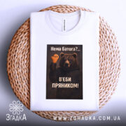 Футболка нема батога. Біла футболка, складена на плетеній кошику, з видимим принтом ведмедя з пряником. Фон білий, що підкреслює деталі принта та текст на українській мові.