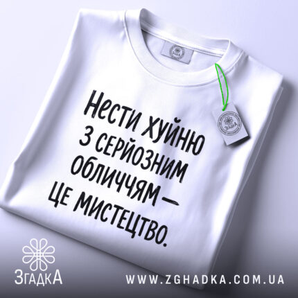 Футболка нести хуйню, білого кольору, з серйозним обличчям, складена на поверхні, фон світлий, принт чорного кольору з текстом: 'Нести хуйню з серйозним обличчям – це мистецтво.', з етикеткою зеленого кольору.