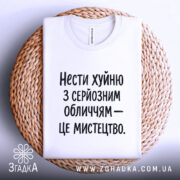 Футболка нести хуйню, білого кольору, складена на плетеній основі, фон світлий, принт чорного кольору з текстом: 'Нести хуйню з серйозним обличчям – це мистецтво.'