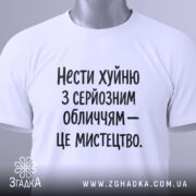 Футболка нести хуйню, білого кольору, з серйозним обличчям, складена на поверхні, фон світлий, принт чорного кольору з текстом: 'Нести хуйню з серйозним обличчям – це мистецтво.', з етикеткою зеленого кольору.