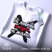 Футболка ні любові, біла, зображена зверху, складена на поверхні, фон світлий, принт чорного кольору з червоними акцентами, текст "Ні любові, ні туги, ні жалю" чітко видимий, на футболці є етикетка з зеленим шнурком.