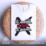 Футболка ні любові, біла, зображена зверху, складена на плетеній основі, фон світлий, принт чорного кольору з червоними акцентами, текст "Ні любові, ні туги, ні жалю" чітко видимий.
