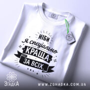 Футболка ніби я краща, білого кольору, складена на поверхні, фон світло-сірий, принт чорного кольору з текстом "Ніби я спеціально краща за всіх" та етикеткою з зеленим шнурком.