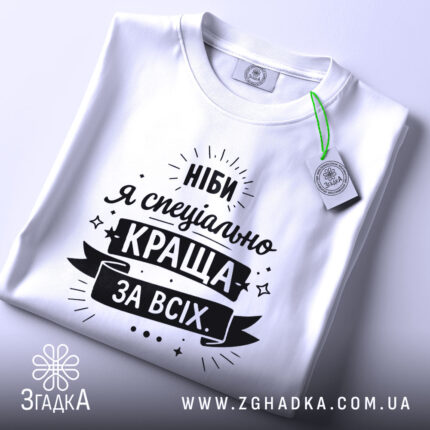 Футболка ніби я краща, білого кольору, складена на поверхні, фон світло-сірий, принт чорного кольору з текстом "Ніби я спеціально краща за всіх" та етикеткою з зеленим шнурком.