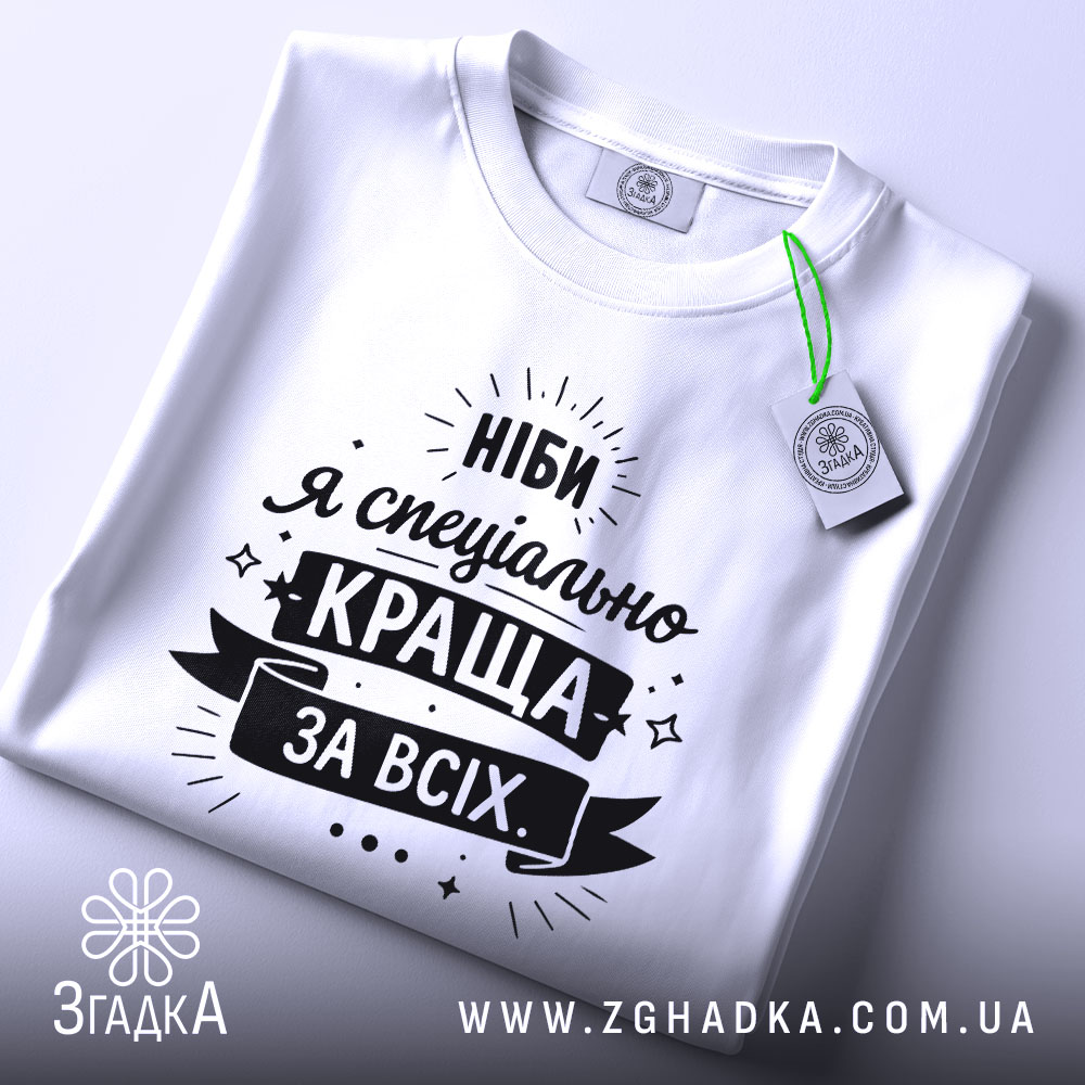 Футболка ніби я краща, білого кольору, складена на поверхні, фон світло-сірий, принт чорного кольору з текстом "Ніби я спеціально краща за всіх" та етикеткою з зеленим шнурком.