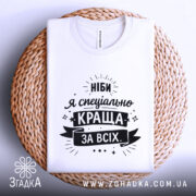 Футболка ніби я краща, білого кольору, складена на плетеній кошику, фон світло-сірий, принт чорного кольору з текстом "Ніби я спеціально краща за всіх".