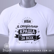 Футболка ніби я краща, білого кольору, складена на поверхні, фон світло-сірий, принт чорного кольору з текстом "Ніби я спеціально краща за всіх" та етикеткою з зеленим шнурком.