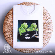 Футболка ніч без сну, біла, з верхнього ракурсу, складена на плетеній основі, з принтом двох зелених ляльок, фон чорний.