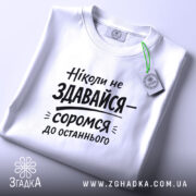 Футболка ніколи не здавайся, білого кольору, складена на поверхні, фон світло-сірий, принт чорного кольору з текстом 'Ніколи не здавайся—соромся до останнього', з етикеткою зеленого кольору.