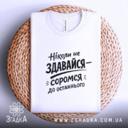 Футболка ніколи не здавайся, білого кольору, складена на плетеній кошику, фон світло-сірий, принт чорного кольору з текстом 'Ніколи не здавайся—соромся до останнього'.