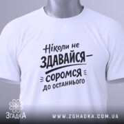 Футболка ніколи не здавайся, білого кольору, складена на поверхні, фон світло-сірий, принт чорного кольору з текстом 'Ніколи не здавайся—соромся до останнього', з етикеткою зеленого кольору.