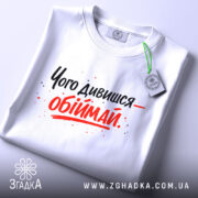 Футболка обійми замість слів. Біла футболка, складена на столі, з принтом чорного та червоного кольорів, текст розташований по центру, на футболці є етикетка з зеленим шнурком.