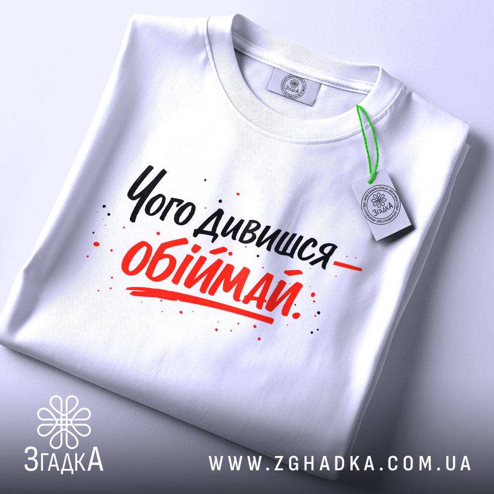 Футболка обійми замість слів. Біла футболка, складена на столі, з принтом чорного та червоного кольорів, текст розташований по центру, на футболці є етикетка з зеленим шнурком.