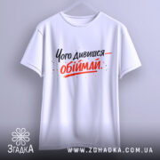 Футболка обійми замість слів. Біла футболка, підвішена на вішалці, з принтом чорного та червоного кольорів, текст розташований по центру, фон світло-сірий.