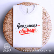 Футболка обійми замість слів. Біла футболка, складена на плетеній основі, з принтом чорного та червоного кольорів, текст розташований по центру, фон світло-сірий.