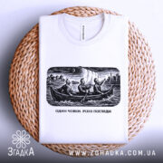 Футболка один човен. Біла футболка, складена на плетеній кошику, з чорно-білим принтом, на якому три чоловіки в човні з вітрилами, фон з міським пейзажем.