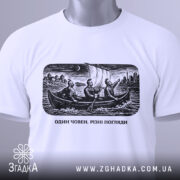 Футболка один човен. Біла футболка, складена на поверхні, з чорно-білим принтом, зображення трьох чоловіків у човні, з вітрилами, на фоні міста, з етикеткою з зеленим шнурком.