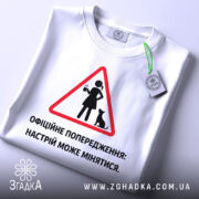 Футболка офіційне попередження: складена біла футболка, зображена зверху, фон світло-сірий, принт у вигляді червоного трикутника з жінкою та котом, текст чорного кольору, на комірці видна етикетка.