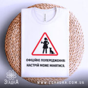 Футболка офіційне попередження: складена біла футболка, зображена зверху на плетеній кошику, фон світлий, принт у вигляді червоного трикутника з жінкою та котом, текст чорного кольору.