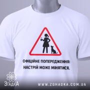 Футболка офіційне попередження: складена біла футболка, зображена зверху, фон світло-сірий, принт у вигляді червоного трикутника з жінкою та котом, текст чорного кольору, на комірці видна етикетка.