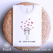 Футболка ой знову ти в голові, білого кольору, складена на плетеному кошику, фон світло-блакитний, принт з малюнком персонажа з сердечками, видно етикетку на комірці.