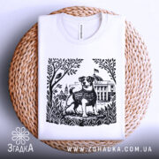 Футболка пес Патрон білого кольору, зображена зверху на плетеній кошику, фон світлий, принт з чорно-білим зображенням собаки Патрона на фоні рослинності та будівлі.