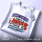 Футболка поруч зі мною, білого кольору, складена на поверхні, принт з червоним дияволом, який читає молитви, фон світло-сірий, видно етикетку з зеленим шнурком.