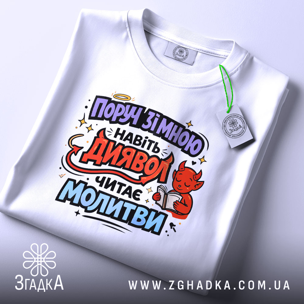 Футболка поруч зі мною, білого кольору, складена на поверхні, принт з червоним дияволом, який читає молитви, фон світло-сірий, видно етикетку з зеленим шнурком.