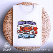 Футболка поруч зі мною, білого кольору, складена на плетеній основі, принт з червоним дияволом, який читає молитви, фон світло-сірий, ракурс зверху.