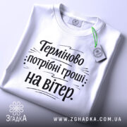 Футболка потрібні гроші. Біла футболка, з круглим вирізом, розташована на столі з легким нахилом. Принт чорного кольору з текстом "Терміново потрібні гроші: на вітер" на білому фоні, з етикеткою, що звисає з коміра.