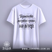 Футболка потрібні гроші. Біла футболка на вішалці, з коротким рукавом, на світлому фоні. Принт чорного кольору з текстом "Терміново потрібні гроші: на вітер" розташований по центру.