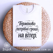 Футболка потрібні гроші. Біла футболка, складена на плетеній кошику, з круглим вирізом. Принт чорного кольору з текстом "Терміново потрібні гроші: на вітер" на білому фоні.