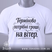 Футболка потрібні гроші. Біла футболка, з круглим вирізом, розташована на столі з легким нахилом. Принт чорного кольору з текстом "Терміново потрібні гроші: на вітер" на білому фоні, з етикеткою, що звисає з коміра.