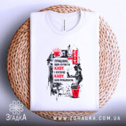 Футболка працюю за каву, білого кольору, зображена зверху на плетеній основі, фон світлий, принт містить текст про каву та малюнок, футболка акуратно складена.