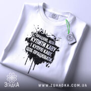 Футболка працюю за каву. Біла футболка, складена на поверхні, з чорним принтом, фон світло-сірий, на футболці є етикетка з зеленим шнурком.