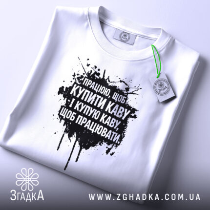 Футболка працюю за каву. Біла футболка, складена на поверхні, з чорним принтом, фон світло-сірий, на футболці є етикетка з зеленим шнурком.