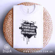 Футболка працюю за каву. Біла футболка, складена на плетеній основі, з чорним принтом, фон білий, принт містить текст з чорними плямами.