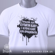Футболка працюю за каву. Біла футболка, складена на поверхні, з чорним принтом, фон світло-сірий, на футболці є етикетка з зеленим шнурком.